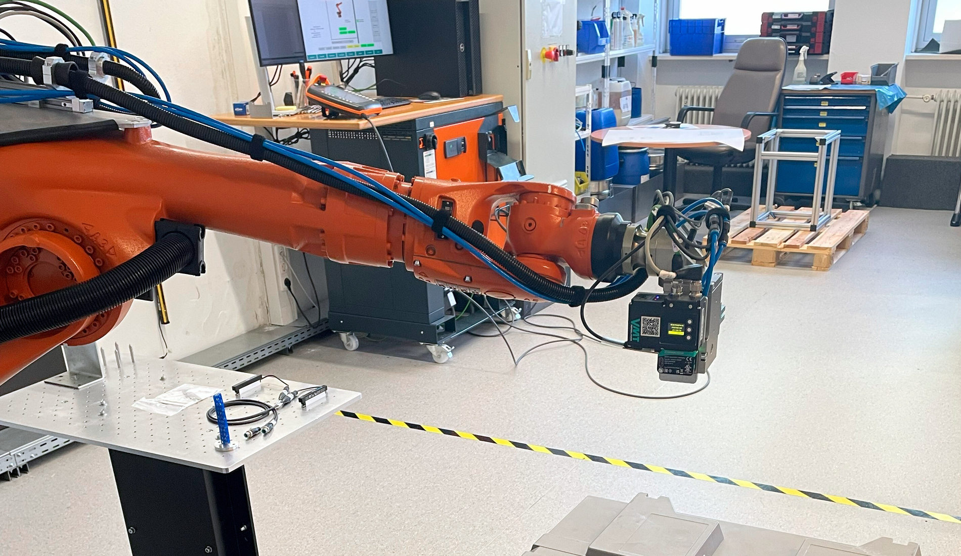 Robotic arm with a gripper in the Polyplan technical lab, with workstations in the background and a teal text panel on the right.